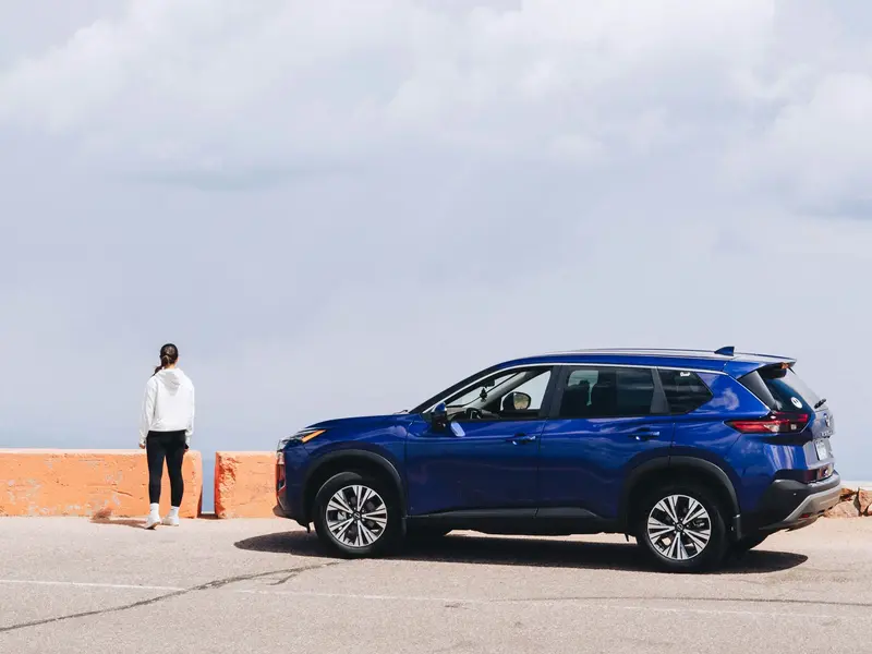 Photo of a blue Nissan Rogue with a woman in front of it looking at the blue sky